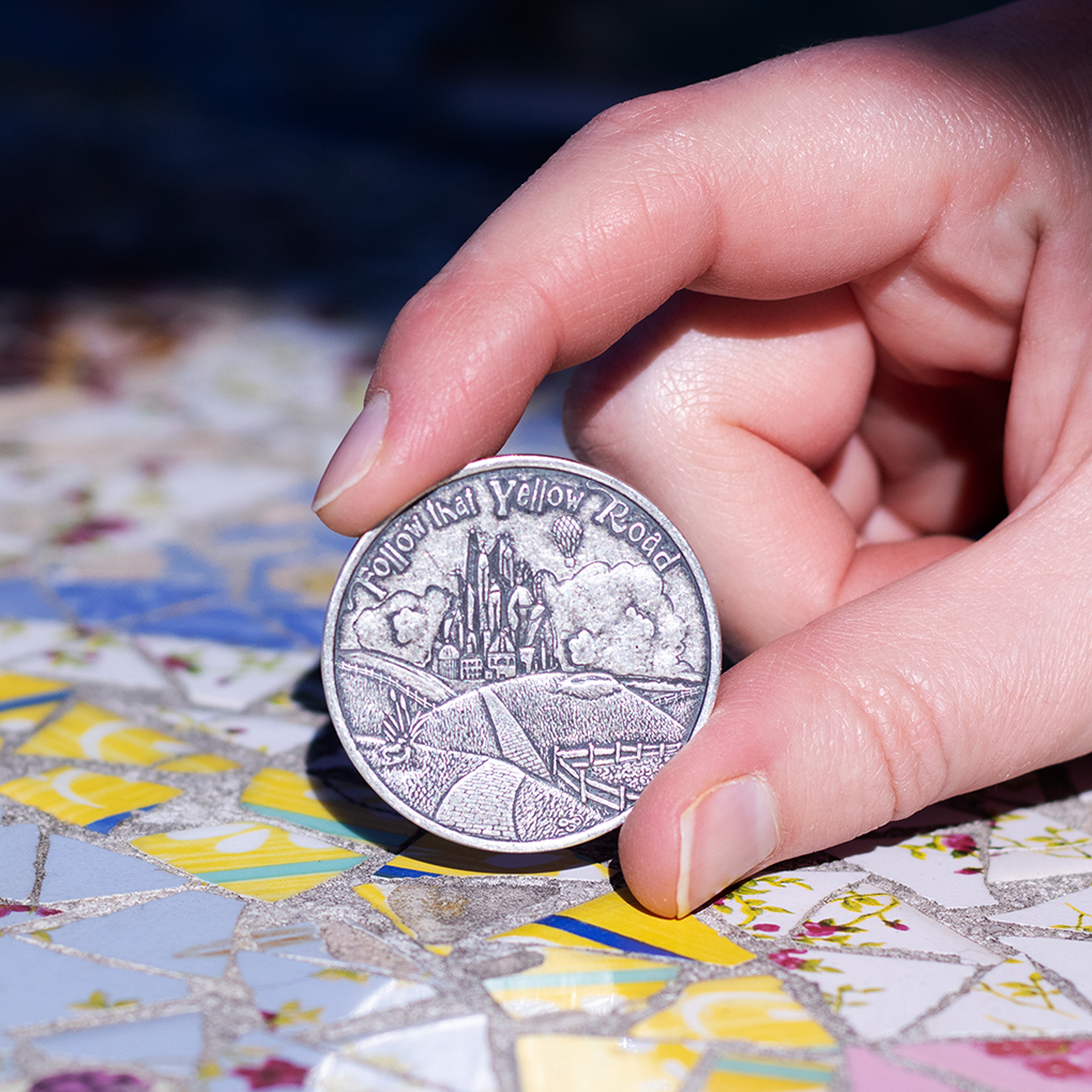 Hand holding silver coin with text, 'Follow That Yellow Road' depicting the Emerald City
