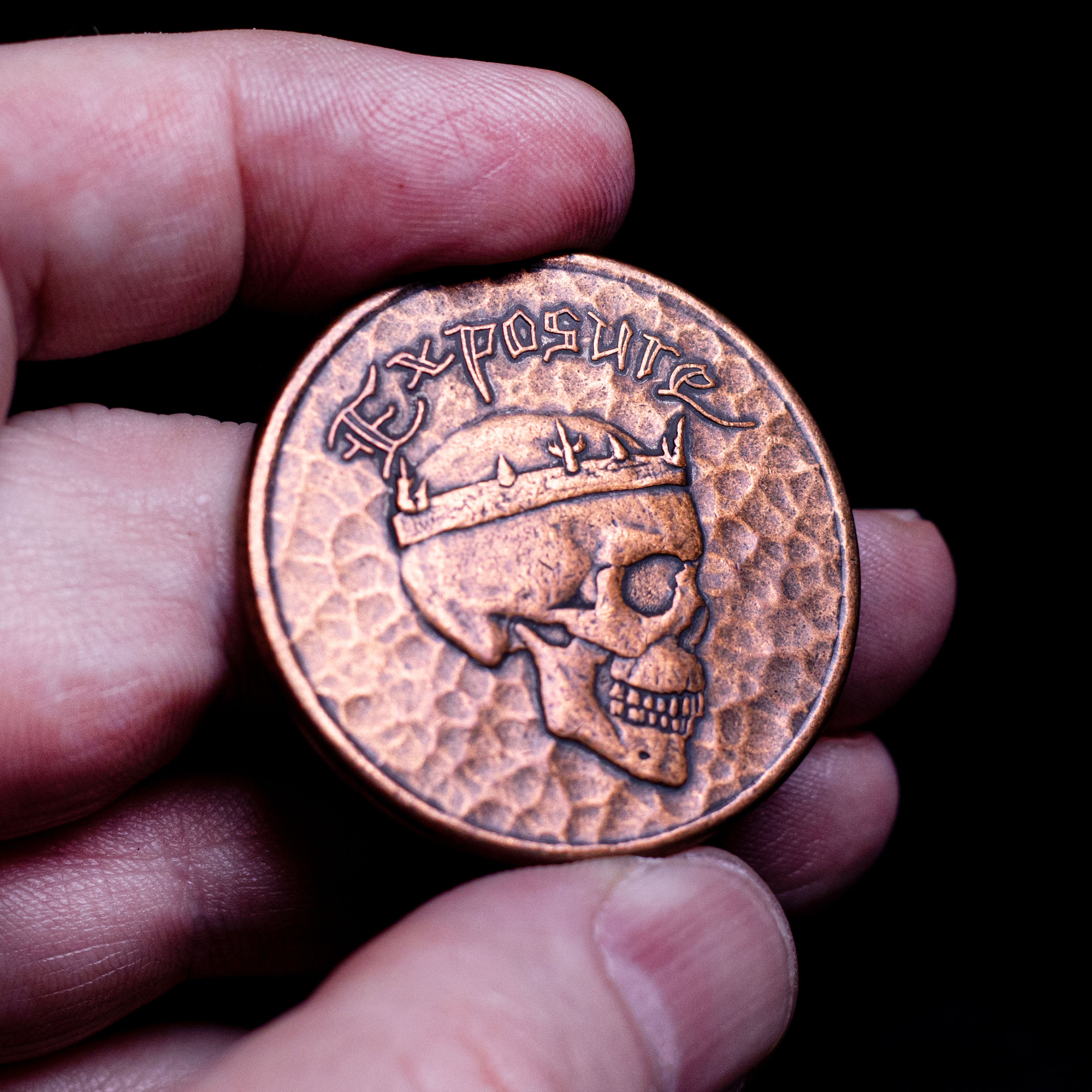 Hand holding copper Exposure Coin, showing the crowned skull