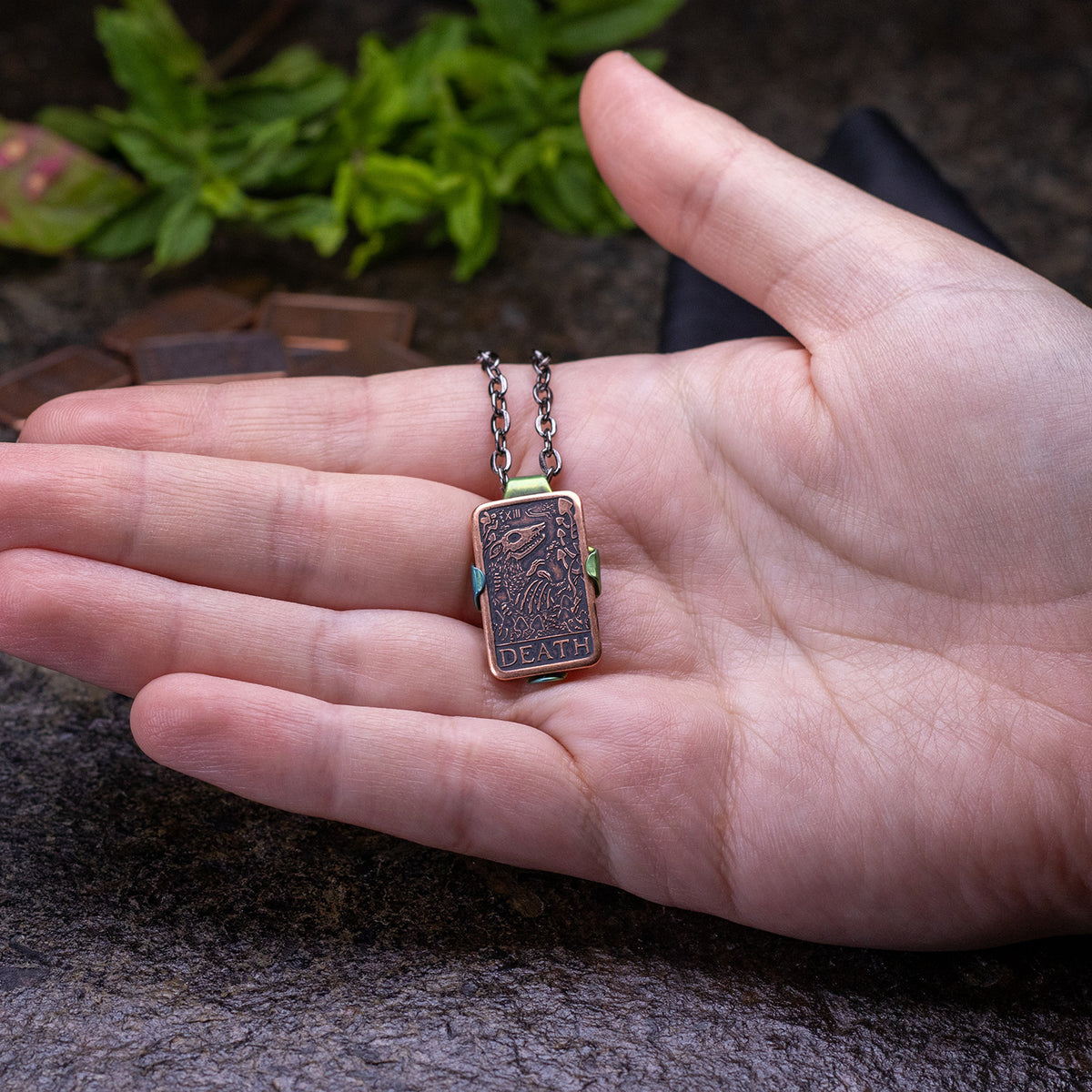Hand holding a niobium, leaf-shaped green pendant holder clasping a copper Tarot Death Coin. 