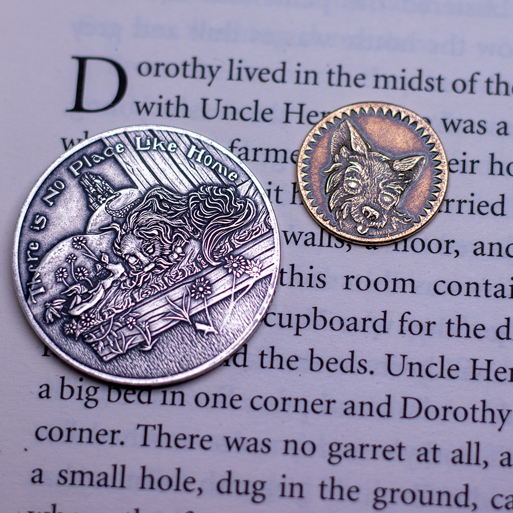 Fine silver coin with text, 'There is No Place Like Home' depicting a sleeping dog on a sunny porch, displayed on a book page next to a bronze penny-size token of Toto from the Wonderful Wizard of Oz