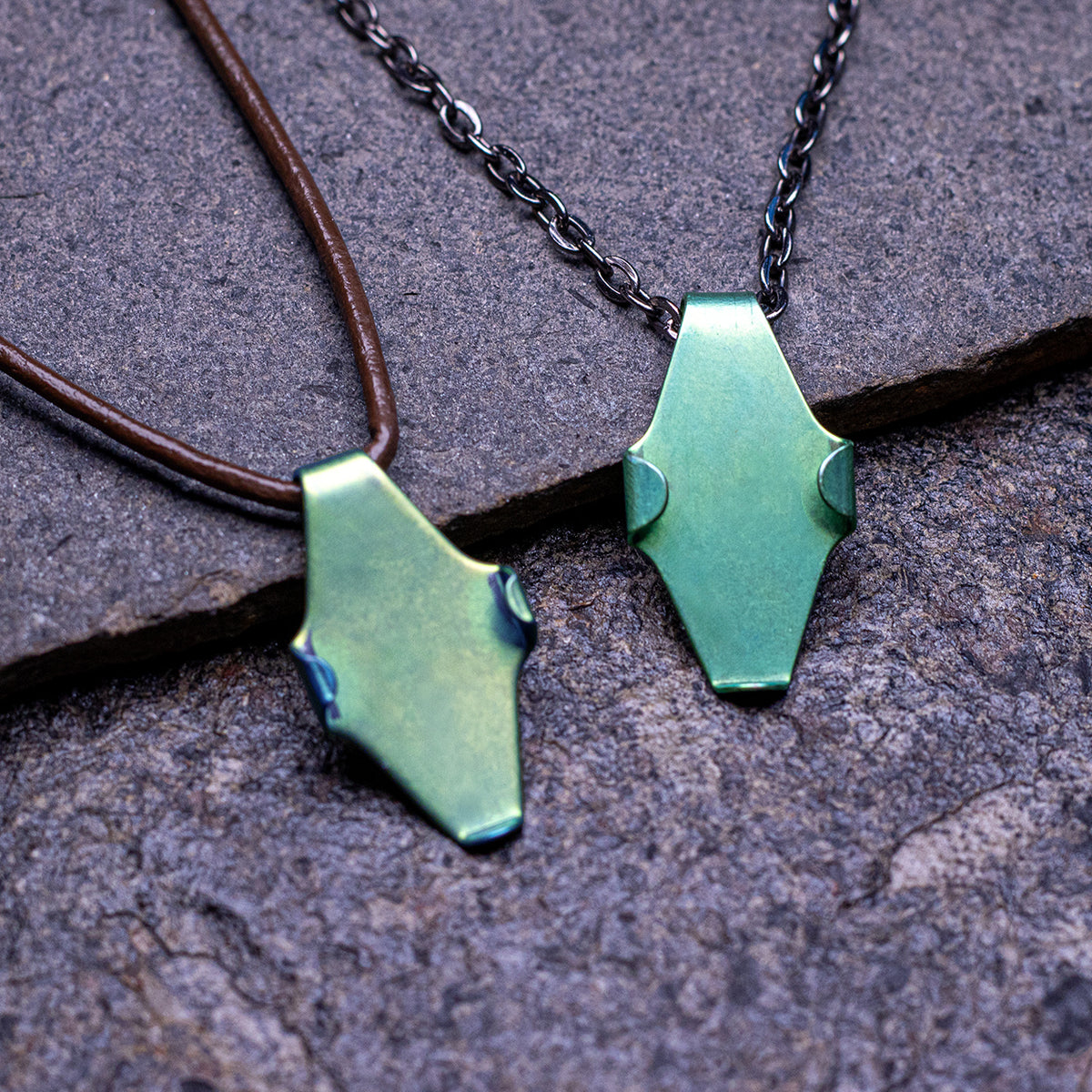 Two leaf=shaped niobium green pendant charms displayed against a rocky background. One has a leather cord, one has a black, steel chain.