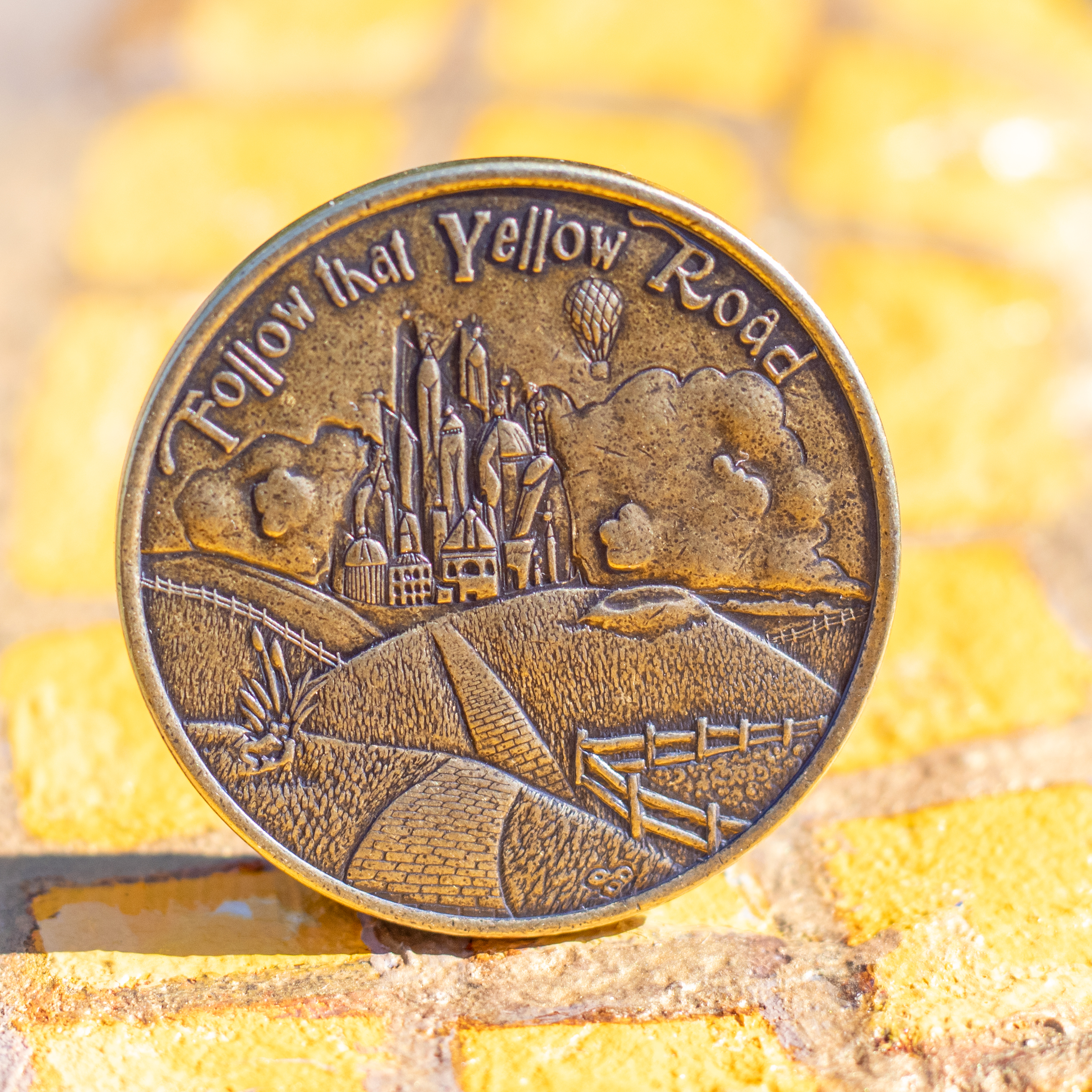 Brass coin featuring the yellow brick road leading up to to the Emerald City, and text 'Follow that Yellow Road'