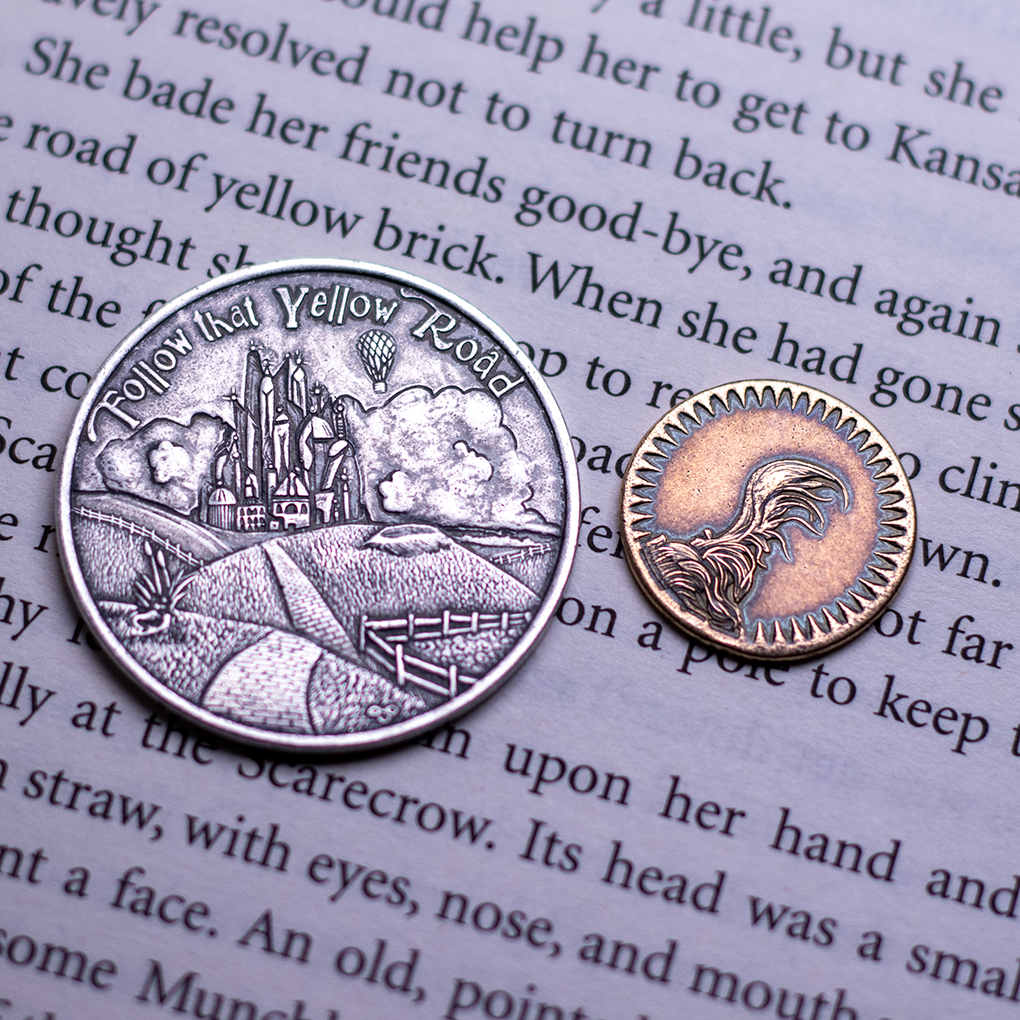 Fine silver coin with text, 'Follow that Yellow Road' depicting the Emerald City and Baum's famous yellow brick road, displayed on a book page next to a penny-size bronze token of a fluffy tail